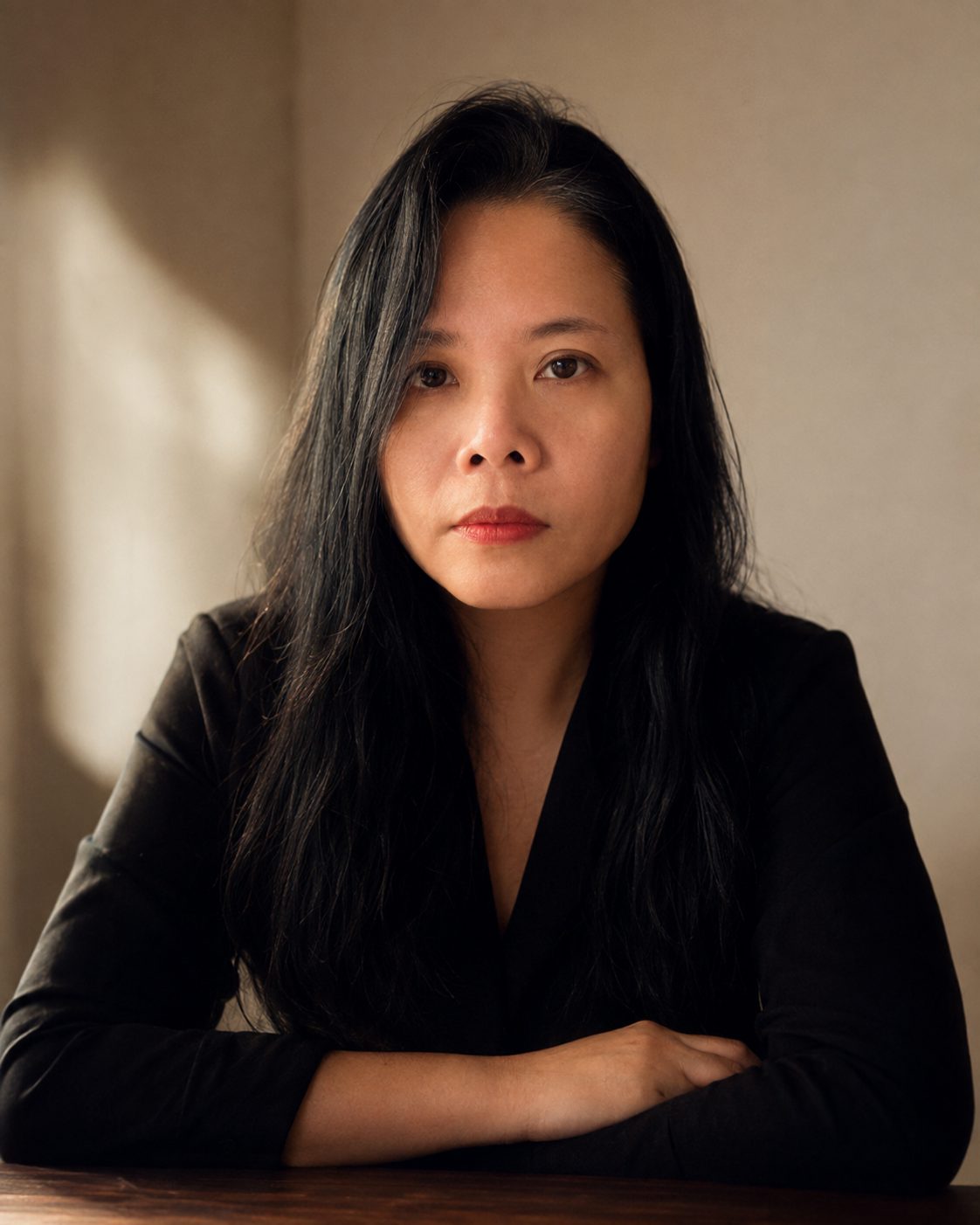 Ayni Chow seated at a wooden table, arms folded, looking directly at the camera in soft window light